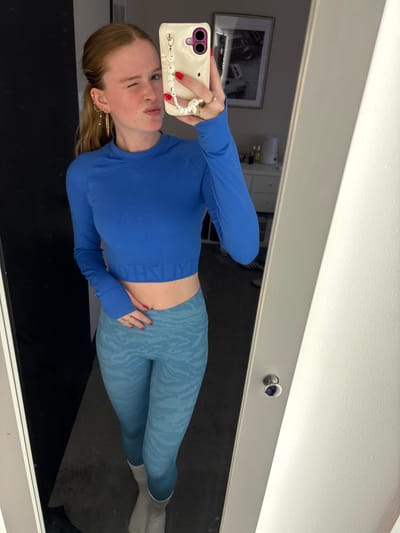 The scene is an indoor mirror selfie depicting a young, fair-skinned female with reddish-blonde hair tied back, located in Rosmalen, Netherlands. She is wearing a royal blue long-sleeved cropped athletic top displaying "GYMSHARK" branding on the lower band, light blue athletic leggings with a subtle pattern, and light-colored ankle socks. Her left hand holds a cream-colored smartphone with a purple camera module and a white beaded wrist strap, pointed towards the mirror to capture her reflection. Her fingernails are painted red, and she wears a ring on her left ring finger and multiple gold-colored earrings in her left ear. She is making a facial expression, winking with her right eye and pouting her lips. The background visible in the mirror's reflection includes a dark floor, a white dresser with various small containers, and a framed black and white photograph of a vehicle mounted on a light-colored wall. The mirror itself has a light-colored frame.