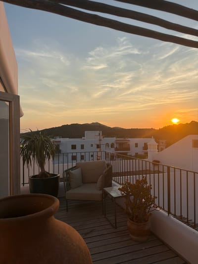 This is a vertical, medium shot of a sunset view from a balcony in Sant Joan de Labritja, Spain. The sun is low on the horizon, casting a warm orange glow across the sky, which is filled with wispy white clouds. In the foreground, there are two large terracotta pots and a wooden railing. Beyond the railing, a white building with several windows is visible, nestled on a hillside covered in green vegetation. A balcony with a chair and small table, as well as a potted plant, are also visible. The overall mood is serene and peaceful, with the sunset creating a beautiful and tranquil atmosphere. The time of day is clearly sunset, and the weather appears to be clear with some clouds.