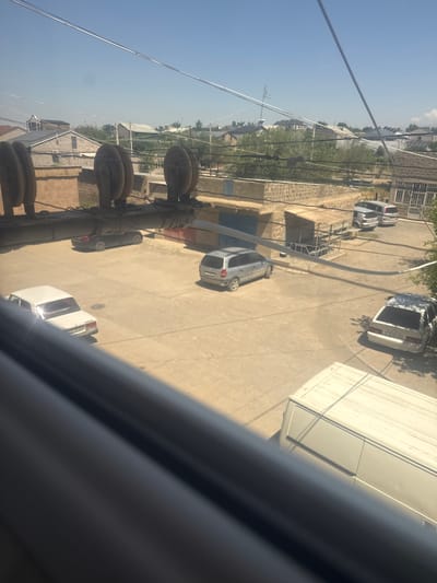 An aerial, eye-level shot looks down on a paved area in the city of Metsamor, Armenia, likely a residential courtyard or an industrial yard. The sky is a clear, pale blue. A few vehicles are parked on the ground: a white sedan, a silver minivan, a dark sedan, and a white panel van. Buildings with brick and stone construction are visible, along with trees and power lines. Three large, wooden pulleys are mounted on a metal bar in the foreground, obscuring the lower part of the scene. The overall impression is one of a quiet, everyday scene in a provincial Armenian city.