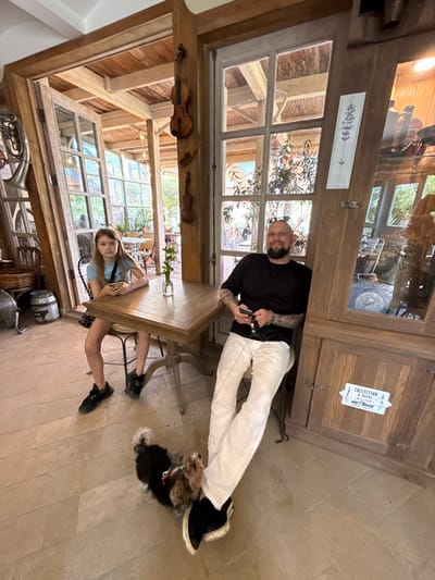 The image depicts a cozy indoor cafe scene, possibly in Semogorskiy, Russia. A young girl with long blonde hair sits at a wooden table, looking at her phone. Opposite her, a man with a shaved head and a beard is seated, also holding a phone. A small dog stands on the floor between them. The cafe has a rustic aesthetic, with exposed wooden beams, glass-paned doors, and decor that includes violins. The floor is made of large stone tiles.