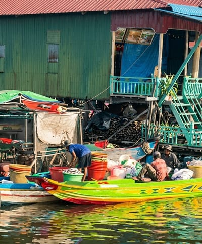 This is a photograph of a floating village in South Nikom, Cambodia. The scene depicts a stilt house made of corrugated metal with a red corrugated metal roof. In front of the stilt house, several boats are docked in the water. One boat, painted green with yellow and red accents, is filled with various items including red and orange buckets, woven baskets, and white sacks. Two men are in this boat; one is wearing a brown shirt and pants and is seated, while the other is standing and appears to be interacting with the contents of the boat. Another boat, partially visible, is painted blue and white. A third boat, primarily visible from the side, is painted green with a red and yellow trim. A man in a blue shirt and black shorts is standing in this boat, attending to some items. The water is a bright, reflective green, suggesting the presence of algae. The overall impression is one of a busy, functional community living on the water.