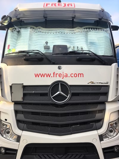 The image shows the front of a white Mercedes-Benz Actros truck parked outdoors, likely in Saint-Augustin-des-Bois, France, given the provided location context. The truck's grille is prominent, featuring the Mercedes-Benz star logo. The windshield is partially covered by a reflective sunshade with decorative tassels. Red lettering on the truck's body reads "www.freja.com". A logo with "FREJA" is visible at the top of the windshield. The sky above appears clear, suggesting daytime. No people are visible in the image, and the truck is stationary. The immediate environment appears to be a paved area, possibly a road or parking lot, with hints of other structures in the background.