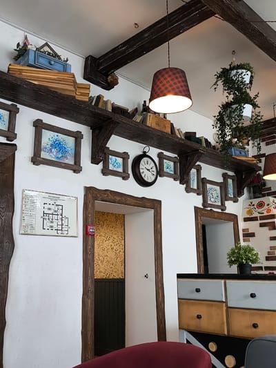 This image captures a warm and rustic interior, likely a cafe or restaurant, characterized by a mix of traditional and repurposed decorative elements. Dark wooden beams crisscross the white ceiling, with a plaid-patterned lampshade hanging centrally, casting a warm light.
A long, dark wooden shelf runs horizontally across the upper wall, adorned with books, a vintage-looking wooden wine box (with visible text "CHATEAU... VIN... DE BORDEAUX"), and a decorative blue wooden drawer containing faux plants. Below this shelf, several framed paintings of blue flowers in various sizes are mounted on the white wall, creating a recurring motif. A large, dark-cased wall clock with Roman numerals is prominently displayed near the center.
On the right side of the frame, several white pots with cascading green foliage hang from the ceiling, adding a natural touch. Further right, a section of the wall features decorative brickwork with vibrant floral patterns.
On the lower left wall, a framed "ПЛАН ЭВАКУАЦИИ" (Evacuation Plan) is visible, with Russian text indicating a "ЛИЦЕЙ И КУЛЬТУРНО-ДОСУГОВЫЙ КОМПЛЕКС" (Lyceum and Cultural and Leisure Complex). Below the plan, a red fire alarm button is mounted. Two doorways, framed with dark, rough-hewn wood, lead to other areas of the establishment. The room through the left doorway features a yellow and green floral wallpaper.
In the foreground on the right, a colorful dresser or cabinet with mismatched drawers in shades of blue, gray, and mustard yellow is partially visible, topped with a small potted plant. A portion of a red chair or stool can be seen in the bottom left corner. No people are present in the scene. The overall ambiance is cozy and inviting, emphasizing a blend of rustic charm and functional design.