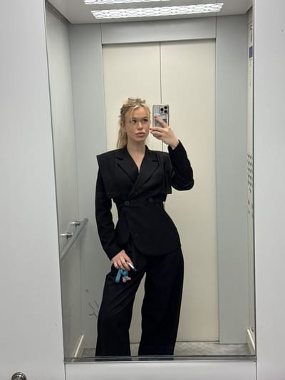 A young woman with blonde hair in a messy bun, wearing a black blazer and matching pants, takes a selfie in an elevator. She is holding a mobile phone in her right hand and a set of keys with a blue charm in her left hand. The elevator walls are metallic grey and white, with a mirrored surface reflecting the woman and the elevator interior. The floor of the elevator appears to be a light-colored tiled surface. There is a silver handrail on the left wall of the elevator. The ceiling of the elevator features a decorative pattern of white squares. This scene is not located in Kommolovo, Russia; it is a generic elevator interior.