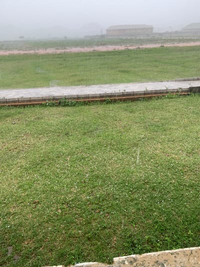 The image depicts an outdoor scene in Farin Yashe, Nigeria, characterized by a hazy atmosphere, likely due to rain or mist. In the foreground, a manicured green lawn is visible, dotted with numerous small, white spherical objects that appear to be hailstones. A paved pathway, bordered by a wooden retaining wall, runs horizontally across the mid-ground, separating the lawn from a red, muddy field beyond. In the distance, several buildings with muted colors are faintly discernible through the haze, along with a lamppost. The overall impression is of a quiet, overcast day with evidence of recent or ongoing hailstorm activity.