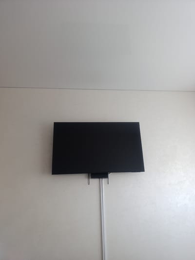 The image shows a black television mounted on a textured, light-colored wall. Above the wall is a white ceiling. A thin, white cable management sleeve runs vertically down the wall from behind the television. A small, dark object is attached to the back of the television, likely a media player or soundbar. The scene is an indoor setting, likely a living room or entertainment area. There are no people or animals visible. The lighting appears to be neutral, suggesting it could be daytime or that artificial lighting is in use. There is no visible text in the image. The location context is Soroca, Moldova.