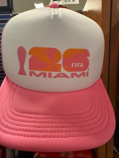 The image shows a pink and white trucker hat. The front of the hat is white with the words "2026 FIFA MIAMI" printed on it in pink and orange. The brim of the hat is pink. The hat is resting on a shelf. The location is Dania Beach, United States of America.