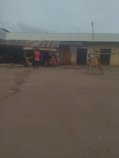 The image shows an empty, dirt street with a few buildings in the background. The street is dirty and dusty, with no people or cars present.  The buildings are made of concrete and have corrugated metal roofs. One building has a sign advertising a product, possibly a brand of soft drink or a food item. The buildings appear to be part of a small market or shop.  There are a few items left on the ground, perhaps remnants from the vendors who had been selling there. The image is taken on a cloudy day, and the overcast sky makes the scene appear dreary. The image is likely taken in Junction, Nigeria, based on the coordinates provided.