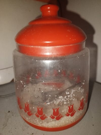 A clear cylindrical plastic jar, featuring a red domed lid and a red base, is centered in the frame. The jar contains white granulated sugar, filling approximately one-third of its volume, with a metallic spoon or scoop partially embedded within. Red tulip designs encircle the lower portion of the jar. The plastic surface is scratched and coated with dust and particulate matter, visible on both the jar and its lid. The object rests on a light-colored, textured surface. The background reveals a light-toned vertical surface, possibly a wall, with a shadow cast behind the jar. This scene is located in Keo Kouang, Laos.