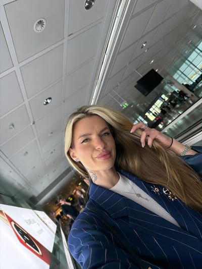 A fair-skinned female subject is depicted in a selfie taken within Munich Airport (MUC) in Freising, Germany. She has long, straight blonde hair extending over her left shoulder, with her left hand lightly touching it. Her face shows defined eyebrows, dark eyeliner, and full, slightly pursed lips. A tattoo resembling a stylized 'S' or snake is visible on the right side of her neck. She is dressed in a navy blue pinstriped blazer worn over a white ribbed top. Accessories include "CD" logo stud earrings, a silver-toned necklace, and two brooches on her blazer's lapel (one appearing as a ship's wheel with a red center, the other as an anchor). Her left hand bears multiple rings on the ring finger, and her left wrist displays a silver-toned metal link watch and a black elastic hair tie. The background reveals a modern airport terminal interior, featuring a white paneled ceiling with recessed lighting and air vents, large glass windows providing a blurred view of an exterior or distant terminal section, a visible escalator or moving walkway on the right, and a suspended black rectangular digital display. A partially visible white advertising stand for "HUAWEI MATE" is present on the lower left. Blurry figures of other individuals are discernible in the deeper background.