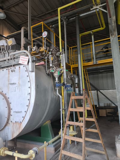 The image shows an industrial boiler room, likely located in Dimona, Israel, given the Hebrew text on a sign. The scene is dominated by a large, cylindrical metal boiler, painted in a light metallic color, with numerous bolts visible along its circumference. Various pipes, some painted yellow and others unpainted metal, are connected to the boiler and extend throughout the space, suggesting a complex network for fluid or gas transfer. A ladder made of light-colored wood is positioned against the boiler, with its rungs clearly visible. Safety railings, painted yellow, are installed at different levels, indicating a multi-story industrial facility. A circular gauge with a red needle is attached to a pipe near the top of the boiler, likely a pressure or temperature indicator. In the background, a desk and an office chair are visible, suggesting a control or monitoring area within the industrial setting. The overall environment appears to be a functional, working industrial space.