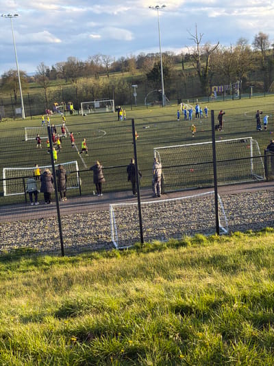 The image shows a sporting ground in Irvinestown, United Kingdom, with multiple fenced artificial turf pitches being used for soccer training. Several groups of children in sports attire are engaged in drills. Adults, presumably coaches or parents, observe from behind the fence. The ground is bordered by trees and grass, and a playground is visible in the background. The sky is partly cloudy.