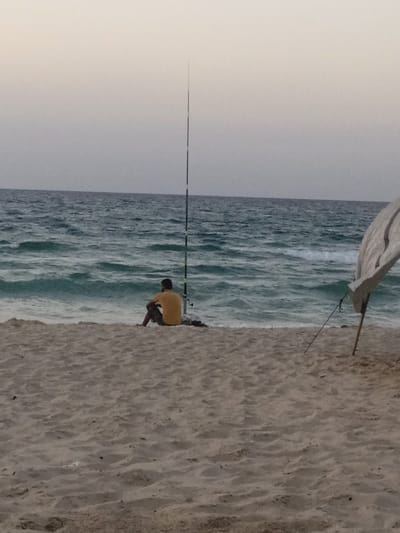 The image depicts a solitary individual engaged in fishing on the beach in Al-Mawasi, Palestinian Territories. In the foreground, a sandy beach occupies the lower third of the frame. In the midground, a person, viewed from behind, is seated directly on the sand, facing the ocean. They are wearing a yellow t-shirt and dark trousers. To the left of the individual, a tall, slender fishing rod is vertically planted in the sand, with its line extending into the wavy, greenish-blue sea. A small dark object or bundle rests on the sand near the fishing rod's base. To the right, a makeshift shelter composed of light-colored fabric or tarp is propped up by two wooden poles. The background features the open sea with moderate waves breaking near the shore, under an overcast or twilight sky transitioning from pale blue to light grey at the horizon.