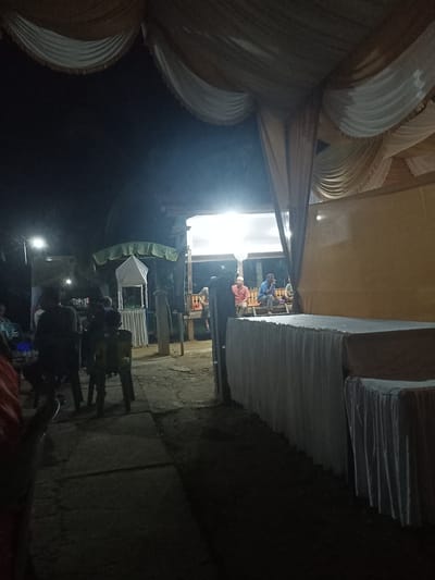 The image depicts an outdoor gathering or event at night, possibly a celebration or a communal meal, in Uleeceubrek, Indonesia. The scene is illuminated by artificial lights, creating a somewhat dimly lit atmosphere. Several people are present, some seated and others standing, appearing to be engaged in conversation or observing something. There are structures that suggest a temporary setup for an event, including draped fabric overhead, possibly from a tent or canopy, and covered tables. In the background, there are structures that look like buildings with lights on, indicating the presence of more established infrastructure. The ground appears to be uneven, possibly dirt or paved with rough materials, consistent with an outdoor location. The overall impression is of a casual and community-oriented event taking place after dark.