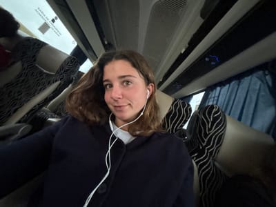 A young adult female is positioned in a medium shot, facing the viewer, inside a passenger transport vehicle, specifically a bus or coach. She has medium-length brown hair and wears a dark blue cardigan over a light-colored top, with white wired earbuds in both ears. Her expression is neutral with direct eye contact. The interior environment features beige seats with a black wavy pattern, dark blue curtains over windows on the right, and an overhead panel with individual lights and vents. A window on the left side displays mirrored Portuguese text reading "SAÍDA DE EMERGÊNCIA" (Emergency Exit). The scene takes place within a vehicle in Cantanhede, Portugal.