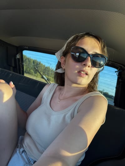 A young Caucasian female is pictured from the chest up, seated in the back of a vehicle. She is wearing a light-colored tank top, denim shorts, and a white bandana tied around her head. Large, dark sunglasses obscure her eyes. A delicate necklace with a cross pendant rests on her collarbone. The interior of the car is visible, with a dark headliner and a textured seat. Sunlight streams in through the rear window, illuminating her arm and casting a shadow across her chest and face. Outside the window, a blur of green trees and a bright blue sky suggests a rural or suburban setting. The image appears to be a selfie, taken with a phone.