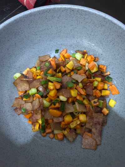 A close-up overhead shot captures a gray, speckled bowl filled with a medley of diced ingredients. The food appears to be a stir-fry or hash, featuring small, uniformly cut pieces of what looks like bacon, orange carrots, yellow bell peppers, and green onions. The bacon pieces are irregularly shaped and slightly curled, with some areas showing a reddish-brown hue. The vegetables are uniformly diced, with the carrots and bell peppers offering vibrant pops of orange and yellow. The green onions are sliced thinly, adding a fresh, green contrast. The ingredients are mixed together and piled loosely in the bowl, with some pieces overlapping. The lighting is warm and slightly dim, casting subtle shadows and highlighting the textures of the food. The background is dark and out of focus, suggesting the bowl is placed on a dark surface. The image does not provide any information or context related to the city of Sukawati, Indonesia.