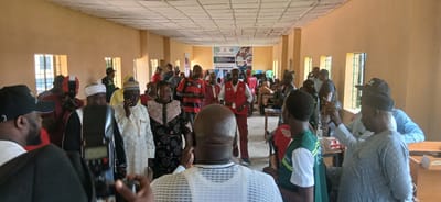 The image depicts an indoor gathering of people in a room with light brown walls and a drop ceiling. The room appears to be a community hall or meeting space, with several windows lining one wall. In the background, a banner is visible with text that reads, "DISTRIBUTION OF EDUCATIONAL MATERIALS FOR LEARNING WITH DISABILITIES IN SABON GARI SOUTH."
The subjects are primarily people of various ages and genders, most of whom are dressed in casual attire or traditional Nigerian clothing. Many individuals are wearing red vests, suggesting they are part of an organizing team or event staff. Some people are holding cameras, indicating that the event is being documented. There are also several tables and chairs set up, with some people seated and others standing and conversing.
The activity taking place appears to be a distribution event, likely related to educational materials for individuals with disabilities. The presence of cameras and the engaged expressions on some faces suggest an active and perhaps noteworthy occasion. The lighting suggests it is daytime, with natural light coming from the windows. The overall atmosphere seems to be one of community engagement and purpose. Visible text on the banner provides specific context about the location and the nature of the event.