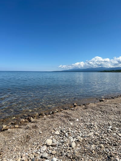 The image shows a wide, tranquil body of water, likely a lake, under a clear blue sky. The water is calm with gentle ripples, and the bottom is visible near the shore, revealing a bed of smooth, rounded pebbles of various sizes and colors. The shoreline is densely covered with these pebbles, creating a natural beach. In the distance, a hazy line of mountains can be seen against the horizon, with some scattered white clouds in the sky. The scene appears to be a natural landscape, devoid of any human presence or artificial structures. Given the context of Tolbazikha, Russia, this is likely a depiction of a shoreline on one of the many lakes in the region.