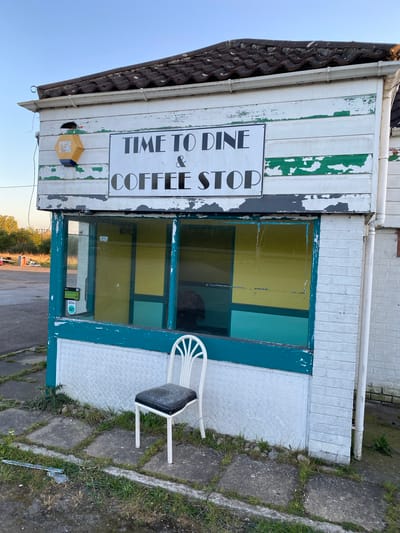 An outdoor, slightly tilted shot captures a weathered, single-story building identified as "TIME TO DINE & COFFEE STOP" in the Breckland District, United Kingdom. The building's exterior features white wooden siding with significant patches of peeling green paint, giving it a distressed look. The roof is covered with dark, ridged tiles, and white guttering is visible along the eaves and down the side.
A prominent rectangular sign on the front of the building reads "TIME TO DINE & COFFEE STOP" in black capital letters on a white background. Below the sign is a large window, composed of several panes, which reflects a mix of yellow and teal hues, likely from the interior or distant elements. To the right of the window, the lower section of the building features a white diamond plate (checker plate) pattern.
An outdoor-style white chair with a black cushion sits on the concrete paving outside the window. The ground consists of cracked concrete slabs with grass growing through the crevices, indicating some neglect. A yellow hexagonal box, possibly an alarm or utility box, is attached to the wall below the main sign. The background, visible through the window's reflection and beyond the immediate structure, suggests a clear sky and possibly a distant road or open field. The lighting implies it is late afternoon or early evening. No people or active events are present, conveying a sense of quiet or abandonment.