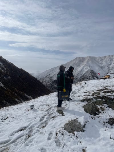 The image depicts two adult males on a snow-covered mountain slope in the mountainous region near the village of Juta, Georgia. The foreground consists of a gentle incline covered by a thin, uneven layer of snow, approximately 5-10 cm deep, through which dry grass and rocks are visible. Multiple footprints are present in the snow.

The first male, positioned on the left and closer to the viewer, is observed from the rear. He is wearing a dark hooded jacket with green stripes on the sleeves, dark pants, and light-colored athletic shoes. He holds a yellow and black DeWalt brand toolbox in his left hand, which is covered by a yellow glove. The second male stands to his right and slightly behind him, viewed from a three-quarter profile. He wears a dark hooded garment over a plaid shirt and has a beard. His hands are positioned near his waist. No overt interaction or specific action is discernible between the two individuals.

The environment includes a dark, vegetated mountain slope dominating the left side of the frame, descending into a distant valley partially obscured by atmospheric haze. To the right, a higher, extensively snow-covered mountain range rises. In the far right background, a small, rustic structure or large tent is visible. Adjacent to this structure, a white flag featuring a large red cross and four smaller red crosses (the Georgian flag) flies on a pole. Two small, dark cylindrical objects, possibly markers or sensors, are partially embedded in the snow on the foreground slope to the left of the first male. The sky overhead is partly cloudy.