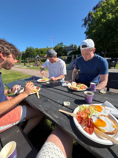 Three young men are eating lunch at a picnic table in Tønsberg, Norway. The table is made of dark wood and is set with plates of food, cups, and napkins. The men are casually dressed, and the weather appears to be warm and sunny. In the background, there is a playground and trees. The man on the left is wearing coral-colored shorts and a blue t-shirt. The man in the middle is wearing a gray baseball cap and a white t-shirt. The man on the right is wearing a white baseball cap and a blue t-shirt. The food on the plates appears to be a breakfast or brunch meal, with eggs, bacon, toast, and salad. The man in the middle is actively eating, while the other two are also engaged with their meals.