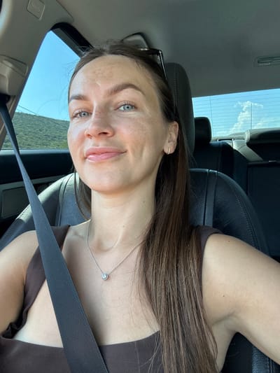 A young woman with blue eyes, light skin, and long brown hair is sitting in the passenger seat of a car. She is wearing a brown top and a silver necklace with a pendant. The car's interior is visible, including leather seats and a seatbelt. Outside the car, a landscape with green hills and a blue sky can be seen. The scene is likely outdoors in a rural or semi-rural area, possibly in Prijeradi, Montenegro.