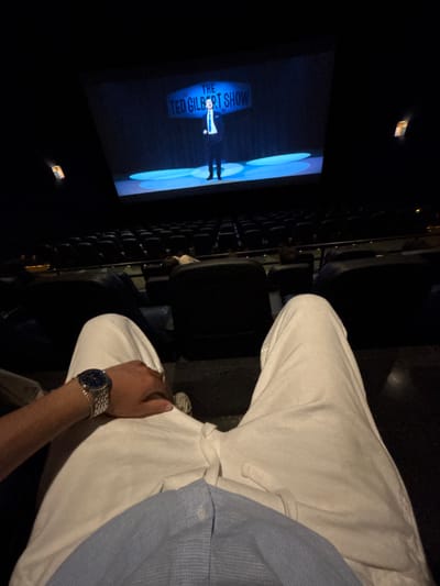 The image captures a seated person in a cinema, viewing "The Ted Gilbert Show" on the screen. The individual wears white pants, a blue shirt, and a silver wristwatch. The scene is dimly lit, typical of a movie theater, with rows of empty seats visible. The image is slightly rotated, providing a unique perspective of the viewing experience.