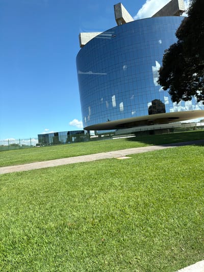 The image captures the Palácio Itamaraty, located in Brasília, Brazil. The building is constructed of a grid-like, mirrored glass facade and supported by concrete pillars. A large green lawn surrounds the palace, bordered by a concrete walkway. The sky is clear and blue.