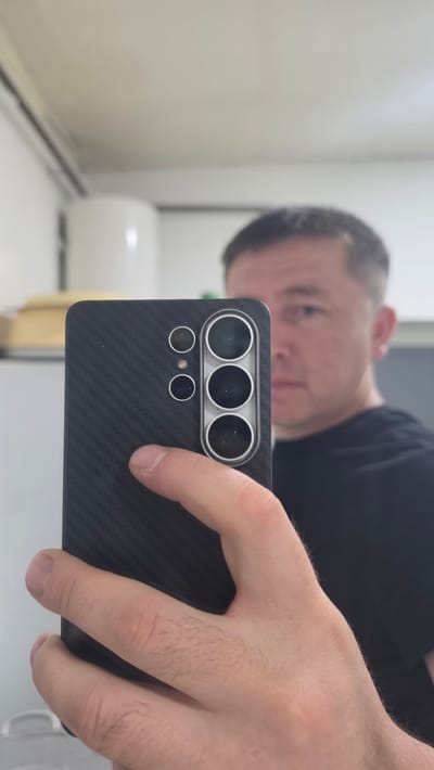 The image is a selfie taken by a person in Kochkor, Kyrgyzstan. The person is holding a smartphone with a black carbon fiber case in their right hand, which is positioned in the foreground, partially obscuring the phone's camera array. The phone's camera module is prominently visible, featuring multiple lenses arranged vertically within a silver housing, along with two smaller circular lenses above. The person's face is visible in the background, out of focus, showing short, dark hair and a black t-shirt. The environment appears to be indoors, with a white cylindrical object and a shelf with boxes visible in the blurred background.