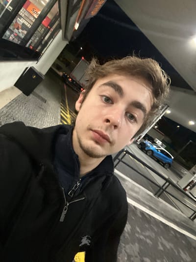 A young man is captured in a close-up selfie at night. He has light brown hair and fair skin. He is wearing a dark jacket with a zipper, possibly standing at a gas station or convenience store. A car and other vehicles can be seen in the background, along with street lights and the store's illuminated windows. The image has a slight tilt.