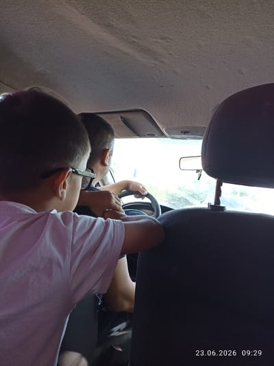 The image shows the interior of a car, with two people visible. In the foreground, a young person wearing a light-colored t-shirt and glasses is seated and appears to be looking out the window or at the driver. Behind and to the right of this person, an adult is seated in the driver's seat, with their hands on the steering wheel. The driver is wearing glasses. The car's interior is visible, including the headrest of the passenger seat in the foreground. The date and time stamp "23.06.2026 09:29" is overlaid on the bottom right of the image. The scene appears to be captured during the daytime, with light coming through the windshield, suggesting the car is in motion or parked outdoors. The location is identified as Batken City, Kyrgyzstan.