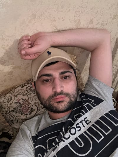The media file is a close-up, high-angle, selfie-style photograph of a man lying down on a textured surface, possibly a bed or couch, with his head resting on a patterned pillow. He is wearing a light-colored baseball cap with a small dark emblem, possibly a logo, on the front and a grey t-shirt with a large black and white graphic design that appears to be text. The text on his shirt is partially visible and reads "LACOSTE". The man has dark hair, a beard, and appears to be looking towards the camera with a neutral or slightly tired expression. His left arm is bent and resting behind his head, with his hand partially visible near his cap. The lighting suggests it could be indoor lighting. The overall mood is relaxed and informal. The location context provided is the Border Gyumri-Akhuryan, Armenia.