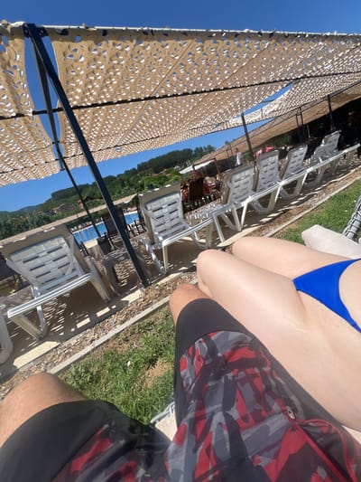 The image captures a sunny day at an outdoor swimming pool facility, likely in or near Delchevo, North Macedonia. In the foreground, two individuals are seated, with one wearing patterned swim trunks and the other a blue bikini. The perspective suggests they are positioned on lounge chairs under a large, patterned shade canopy. Rows of empty white plastic lounge chairs are visible, extending towards the pool area. The pool itself, filled with bright blue water, is visible in the background, along with a treed hillside dotted with houses. The overall environment is recreational, indicating a vacation or leisure setting.