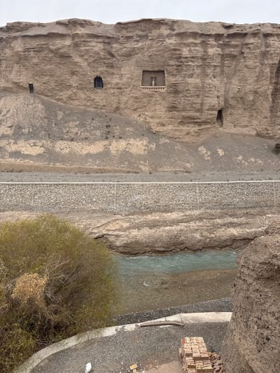 The image shows a vertical view of a scene at the Yulin Caves in Guazhou County, China. On the left, a large cliff face with cave openings and constructed windows is visible. A paved pathway runs along the base of the cliff. To the right of the path, there's a river or stream. Further to the right, a stack of bricks is visible near the edge of the image. Trees and gravel are also present in the scene.