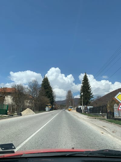 The image captures a rural road scene in Gintsi, Bulgaria, viewed from inside a moving vehicle. The asphalt road stretches into the distance, marked by a prominent solid white line on its right side. On the left of the road is a dirt or gravel shoulder, beyond which are various trees and buildings.
The sky is a clear, vibrant blue, adorned with large, fluffy white cumulus clouds. Multiple power lines traverse the upper portion of the sky. Along the roadside, there's a mix of vegetation, including tall evergreen trees with full, dark green foliage and several deciduous trees that are currently bare, suggesting the season is late winter or early spring. Behind the trees, several houses with characteristic reddish-brown tiled roofs and fences are partially visible.
Further down the road on the left, a yellow construction vehicle, appearing to be an excavator or similar heavy equipment, is parked or positioned near the roadside, accompanied by a smaller white utility vehicle. In the upper left quadrant of the image, a portion of a yellow diamond-shaped road sign with a black border, typically indicating a priority road, is visible. Another road sign is partially obscured next to it. The entire scene is bathed in bright daylight, indicating a sunny day. The right edge of the frame shows elements of the vehicle from which the photograph was taken, including a red painted surface and a black panel with some small, illegible white marks. No explicit text is clearly discernible on any signs or objects.