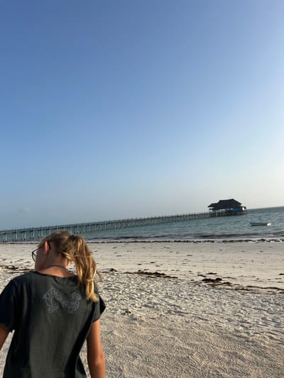 This vertical image captures a serene daytime scene on a beach in Kiwengwa, Tanzania. The dominant feature on the left is a vast, clear blue sky, graduating slightly in tone. On the right, a sandy beach meets calm, blue-green ocean waters.
In the foreground, a person is visible from the back and side, looking downwards. They have blonde hair tied in a ponytail and are wearing glasses and a dark t-shirt. On the back of the t-shirt, a light-colored graphic depicts two stylized marine creatures, possibly fish or shrimp, arranged symmetrically.
The beach itself is composed of light-colored sand, with scattered patches of darker seaweed or debris. A long, narrow wooden pier extends from the shore into the ocean. At the far end of this pier, an overwater structure with a dark, peaked roof stands out against the water. A small, dark boat is also seen floating in the water near the structure and the end of the pier.
The weather appears clear and sunny, suggesting a pleasant day. The setting evokes a tropical coastal environment. No specific text is legible within the image beyond the stylized graphic on the person's shirt.
