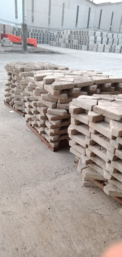 The image shows stacks of rectangular paving stones on wooden pallets. The stones are light beige and appear dusty or weathered. In the background, there is a large building with tall, narrow windows and stacks of U-shaped concrete structures. An orange tarp partially covers some of the structures in the background. The location is Sumput, Indonesia, likely an industrial or storage area. There are no people visible. The focus is on the paving stones and the materials yard environment.