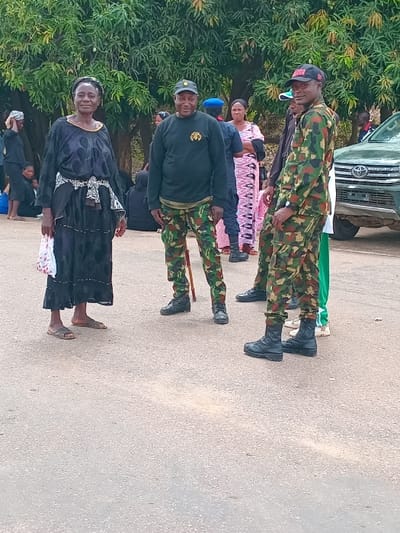 The scene is a paved road in Ungwan Tanko, Nigeria (latitude: 10.422, longitude: 7.49), where a group of people are gathered. There are six individuals in the scene. A woman in black clothing is standing in the foreground, facing the camera. She is holding a white and red bag in her left hand. To her right, a man in black clothing and camouflage pants is standing with a cane. Behind him, a man in a green and camouflage uniform is standing with his arms crossed. To the right of him, a man wearing a black and white checkered shirt and a woman in a pink and black dress, are standing together, both looking at the camera. In the background, there is a military vehicle parked on the right side of the road. There are some other people standing and sitting in the background, blurred out of focus. The scene is outdoor, with lush foliage in the background. The sky is not visible.