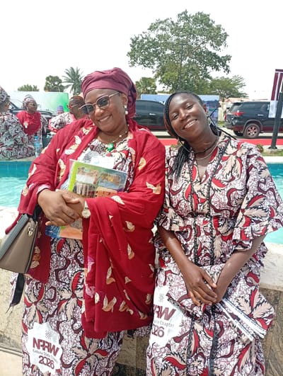 Two women are prominently featured in a bright outdoor photograph, likely taken at an event in Unguwan Sanusi, Nigeria. The woman on the left is smiling broadly, wearing sunglasses, a red headwrap, and a flowing red kaftan adorned with gold leaf-like patterns. She holds a stack of books or documents with colorful covers and wears a patterned dress underneath her kaftan. Her accessories include a necklace, earrings, and a wristwatch, with a dark handbag visible on her left.

Beside her, the woman on the right is also smiling, dressed in a garment with a matching red, beige, and black patterned fabric, which appears to be the same textile as the inner dress of the woman on the left. She has braided hair and wears necklaces and large hoop earrings. Both women are seated next to a blue-tiled water feature, possibly a fountain or pool.

In the background, other women in similar attire can be seen, along with dark SUVs, lush green trees, and distant structures, indicating an outdoor gathering or event. The overall atmosphere appears joyful and social, conveyed by the subjects' expressions.

Visible text on the patterned fabric includes "NPRW Kaduna State Chapter 2026" and a textile brand label that reads "VERITABLE BLOCK PRINTS NEW CHIGAN". These details suggest the event is associated with the "NPRW Kaduna State Chapter" and that the traditional attire is made from a specific type of printed fabric.