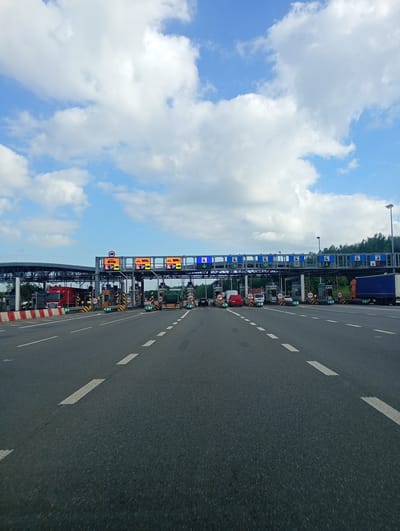 The image shows a multi-lane toll plaza on a highway. Several vehicles, including trucks, vans, and cars, are visible at the toll booths. Overhead gantries display traffic signs and lane indications. The sky is blue with scattered white clouds. The foreground shows the asphalt road with white lane markings. Based on the context provided by the query, this toll plaza is located in Mysłowice, Poland.