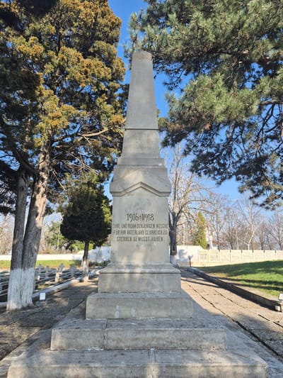 The image depicts a World War I memorial in a cemetery setting. The inscription reads "1916-1918, EHRE UND RUHM DERJENIGEN WELCHE FUR IHR VATERLAND GLORREICH ZU STERBEN GEWUSST HABEN." which translates to "1916-1918, HONOR AND GLORY TO THOSE WHO KNEW HOW TO DIE GLORIOUSLY FOR THEIR FATHERLAND." The memorial is made of stone and stands amidst trees and other grave markers. It is located in Shumen, Bulgaria.