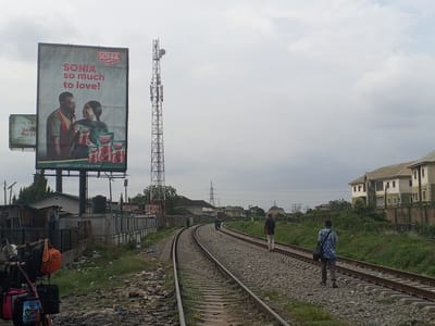 The image depicts a railroad track in what appears to be Jibowu, Nigeria. Several individuals are walking along the tracks. A large advertisement billboard for 'SONIA' tomato paste stands prominently next to the tracks. A tall cell tower is visible behind the billboard. Buildings and overgrown vegetation are present on either side of the tracks. The sky is overcast. A luggage carrier with bags is present on the left side.