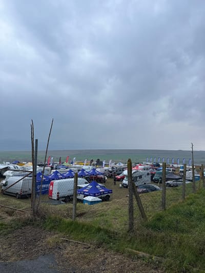An outdoor scene in Poroy, Bulgaria, captured under a uniformly overcast, light-grey sky. The foreground features a wire mesh fence with concrete posts, extending across the bottom half of the frame. Two slender, vertical bare branches or stakes rise behind the left portion of the fence. The ground in the immediate foreground consists of dry, sparse grass and bare earth.

Beyond the fence, a large, open grassy field hosts an extensive, temporary encampment. This area is densely populated with numerous vehicles, primarily white cargo vans, assorted recreational vehicles (RVs) and camper vans, and several pickup trucks, including a red model. A multitude of pop-up canopies, predominantly blue with some white and red examples, are erected throughout the field. Visible branding on these canopies and accompanying flags includes "YAMAHA," "MS Lubricants," and "Liqui Moly." Tall, vertical advertising flags, largely blue and white, are arranged in rows and clusters within the setup. At least two individuals are discernible in the midground among the vehicles and tents, appearing to be standing. A blue portable toilet or utility box is present under a blue canopy.

The background reveals distant, low-lying hills or plains under the same grey sky, indicating a vast, open landscape beyond the event area. The overall impression is that of a motorsports or outdoor rally staging ground or event site.