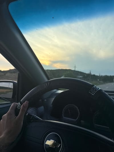 The image shows the interior of a car, viewed from the driver's perspective. A hand is visible on the steering wheel, which is covered in a black case adorned with silver studs. The dashboard and steering wheel are dark, with a Chevrolet logo visible on the latter. Through the windshield, a sunset is seen with a gradient of blue and yellow sky. The landscape outside appears rural, with hills and scattered vegetation. The location is specified as the city of Sadaclia, Moldova, though this information cannot be visually confirmed from the image itself. The overall impression is that of a person driving at dusk in a semi-rural area.