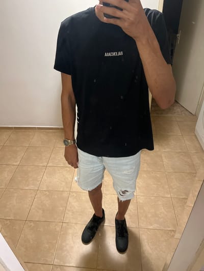 A person is standing in what appears to be a residential hallway or room with tiled flooring. The person is wearing a black t-shirt with white text, light blue distressed denim shorts, and black sneakers. A gold watch is visible on the person's left wrist. The background shows a plain white wall and a doorway. There are no identifiable landmarks or clues suggesting the location is specifically in the city of Voysil, Bulgaria.