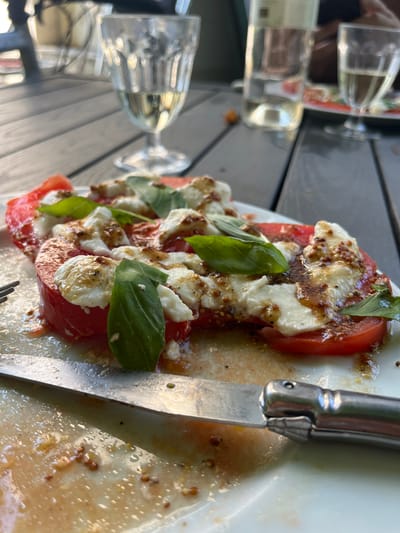 This close-up, slightly angled shot showcases a Caprese salad on a white plate. Slices of ripe red tomatoes are layered with chunks of fresh mozzarella cheese and garnished with vibrant green basil leaves. The salad is generously drizzled with a dressing that appears to be vinaigrette with whole grain mustard seeds, creating a visually appealing texture and shine. A knife with a metallic handle rests on the right side of the plate, positioned as if ready for use. In the background, a wooden table surface is visible, along with glimpses of glassware and what might be a bottle, suggesting an outdoor dining setting. The lighting suggests it is daytime, possibly late afternoon given the soft light. The overall impression is one of a fresh and appetizing meal, likely enjoyed at a restaurant or a casual outdoor meal.