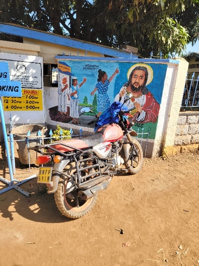This outdoor image, taken in bright daylight under a clear blue sky, captures a vibrant mural on the wall of a building in Wang'uru, Kenya. The ground in the foreground is unpaved dirt, and lush green foliage from trees is visible on the left side of the frame.

The central feature is a large, colorful mural painted on the white upper section of the building. It depicts Jesus Christ at the top, with a halo, long brown hair, and a beard, wearing a red robe and holding a lamb. Below him, several figures are painted, including a woman in a blue polka-dotted dress with her arms raised, and two children, one standing and one sitting. Green trees and a landscape form the background of the mural. Partial text on the mural reads "AW HEALDO" and "ALS".

Parked directly in front of the mural is a red and white motorcycle, oriented sideways, facing to the right. It features a robust cargo rack on its rear and a visible Kenyan license plate, "KMFP 691M". A light blue metal fence or barrier is partially visible in the immediate foreground, obscuring the motorcycle's front wheel.

Below the mural, on the lower, yellowish section of the building, two signs are mounted. A larger white rectangular sign, partially obscured, shows a logo and some text, including "FROTARY CERIN M" and "OKING". Adjacent to it, a smaller yellow sign displays "HOURS:" followed by specific time slots: "07-00AM", "12.30PM - 2:00PM", and "4.00PM - 5-00PM". The scene suggests a community building or center, possibly with religious or social services.
