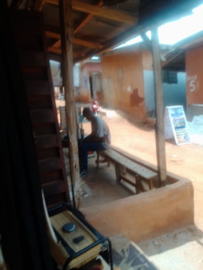 The image shows a man sitting on a wooden bench under a corrugated metal roof, supported by wooden pillars, in the town of Auchi, Nigeria, at the coordinates (latitude: 7.062, longitude: 6.273). The man is wearing a light-colored shirt and blue jeans. He is sitting with his head down, appearing to be engaged in a task. The bench is made of rough planks of wood and is set on a dirt floor. There is a small generator visible in the bottom left corner of the image, partially obscured by a dark curtain. The scene suggests a simple, outdoor setting, possibly a workshop or small business. The background is a dirt alley with a building with a brown brick wall, visible on the right. The building has a sign with a number "5" on it. The image is taken from a slightly elevated angle, looking into the shaded area under the roof. The light is bright and slightly harsh, likely midday sun. The scene is relatively clean and well-maintained.