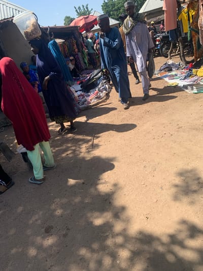 This is a medium shot, taken from eye-level, of a busy outdoor market scene in Boi A, Nigeria. The ground is a dusty, sandy dirt path, and the scene is dappled with shadows from overhead trees or structures.

In the foreground left, a person wearing a bright red hijab and light green pants walks away from the camera. To their right, a woman in a dark blue hijab and dress also walks away, carrying a bucket on her head filled with food items. Behind her, another woman in a blue headscarf is visible, looking towards the market.

The middle ground is dominated by market stalls. One stall has a variety of colorful textiles and clothing for sale, with mannequins or displays of garments. Another stall appears to be selling food or goods in containers. Several people are browsing or working at the stalls. A man in a light blue robe and cap walks towards the right of the frame, and another man in a similar light blue robe and a patterned scarf is walking next to him. A motorbike is parked near one of the stalls.

In the background, there are simple buildings with corrugated metal roofs, typical of the area. The sky is bright and clear, indicating a sunny day. The overall impression is one of vibrant activity and commerce in a rural Nigerian market.